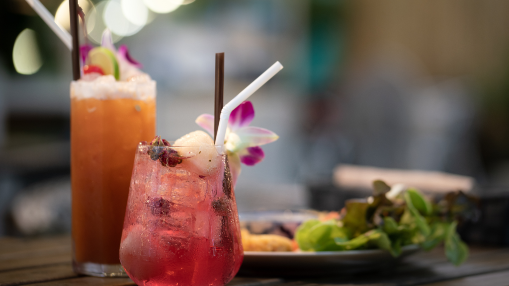 Close-up of two refreshing mocktails with ice, garnished with flowers and fruit, served on a wooden table with a salad plate.